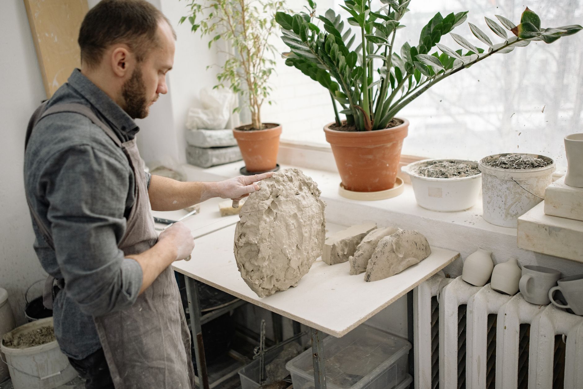 A focused potter sculpting clay on a table in a well-lit indoor studio with plants.