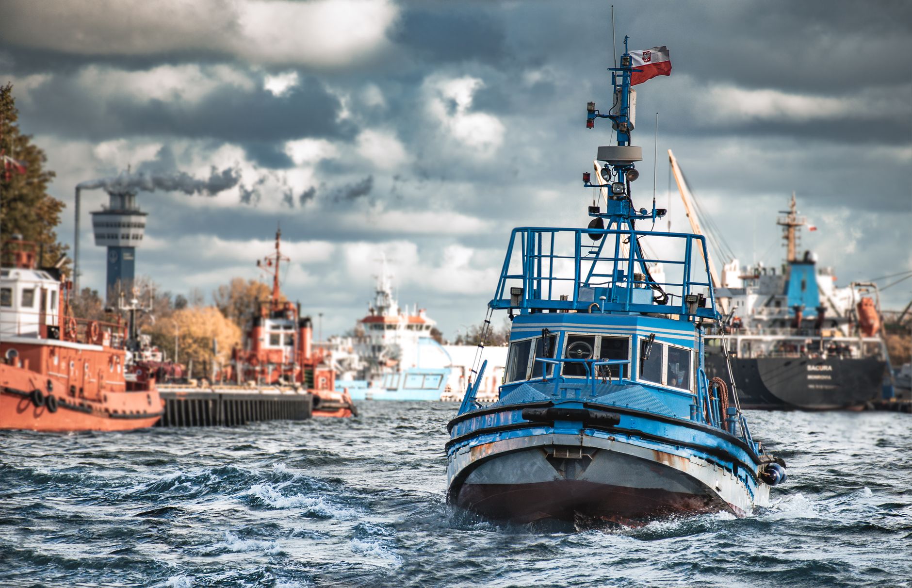 A blue tugboat sails through the busy Gdańsk shipyard with industrial background.