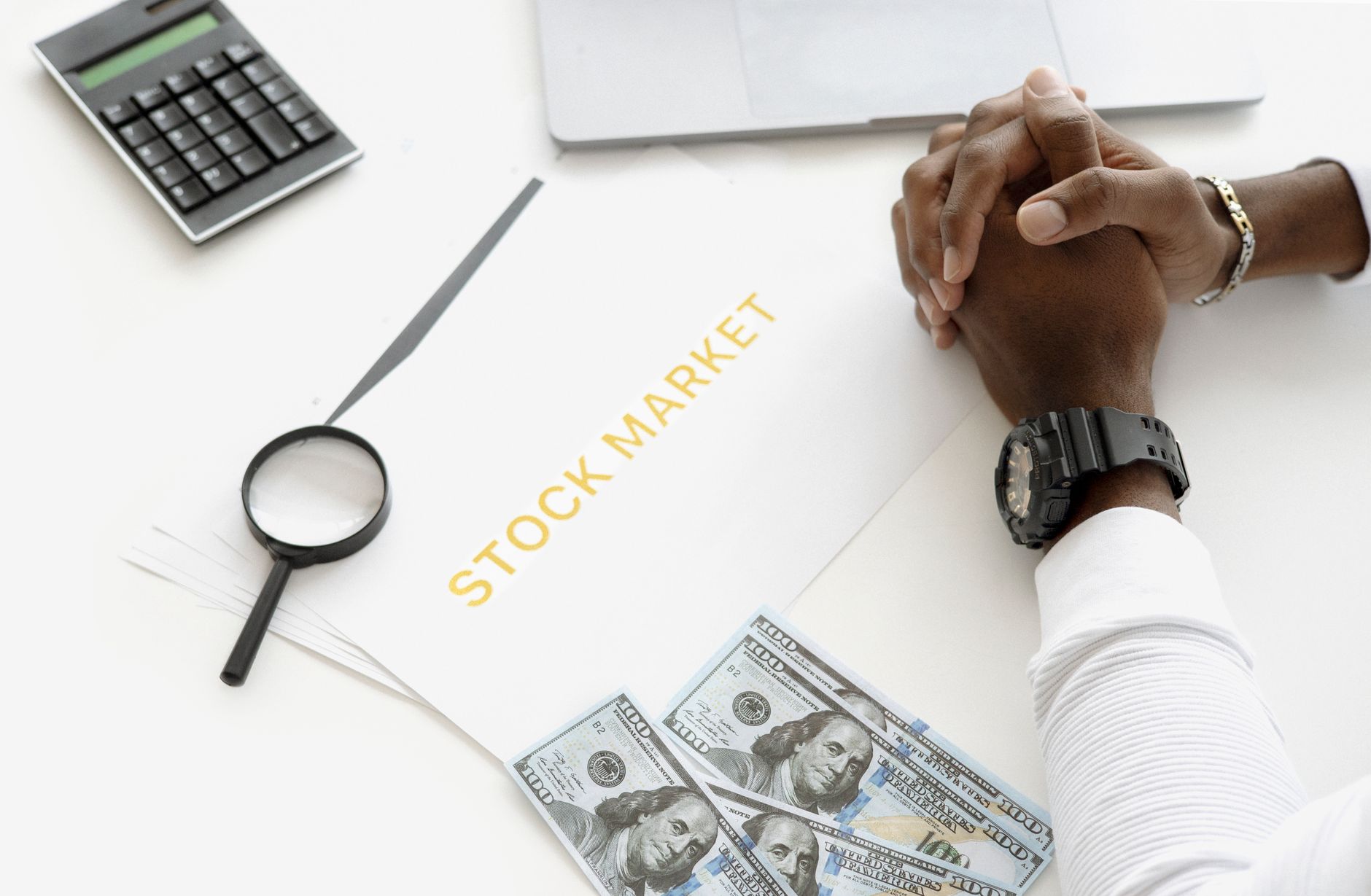 Hands folded over stock market documents with calculator and cash on desk.