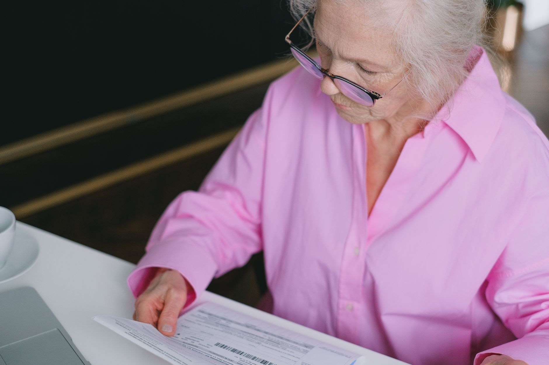 Elderly woman in pink shirt carefully reading a document indoors.