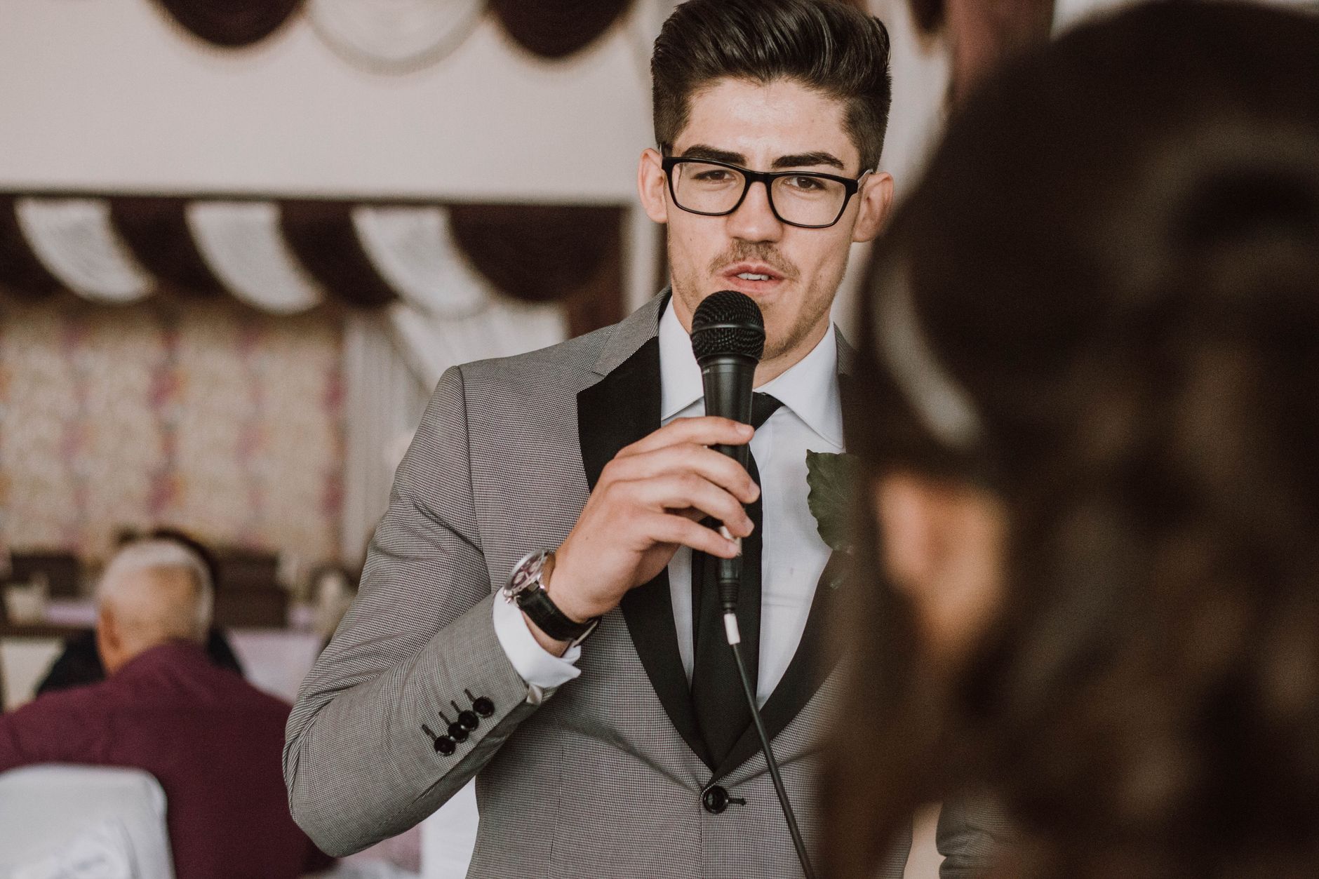 A man in a suit and glasses speaking into a microphone at a formal indoor event.