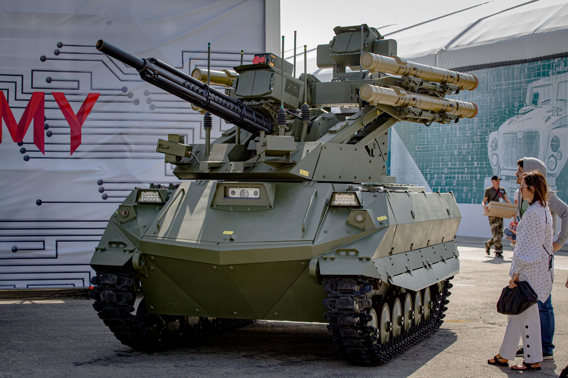 Green military tank with missile launchers on display outdoors with spectators.