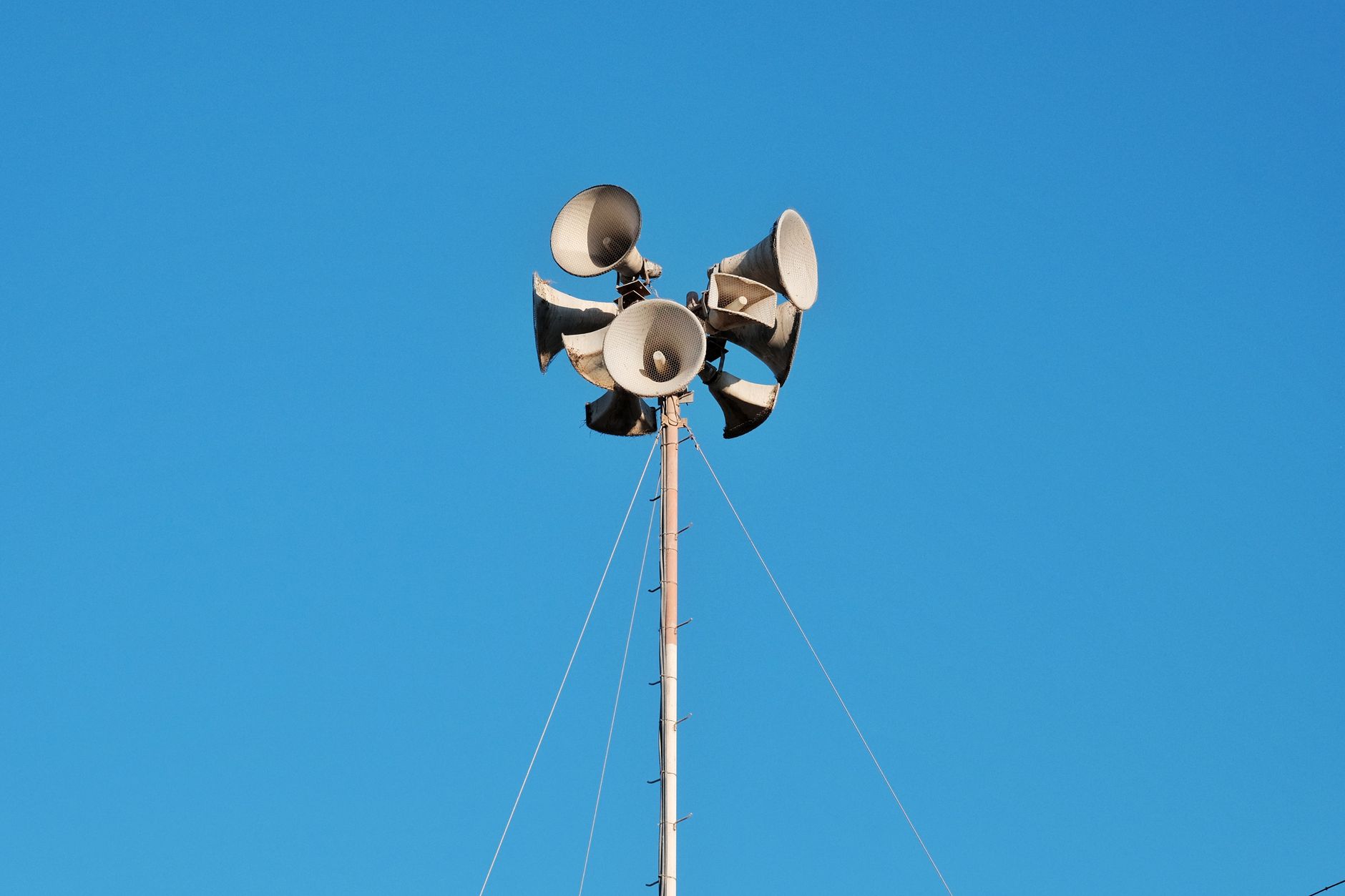 High pole with multiple loudspeakers against a vibrant blue sky, urban setting.