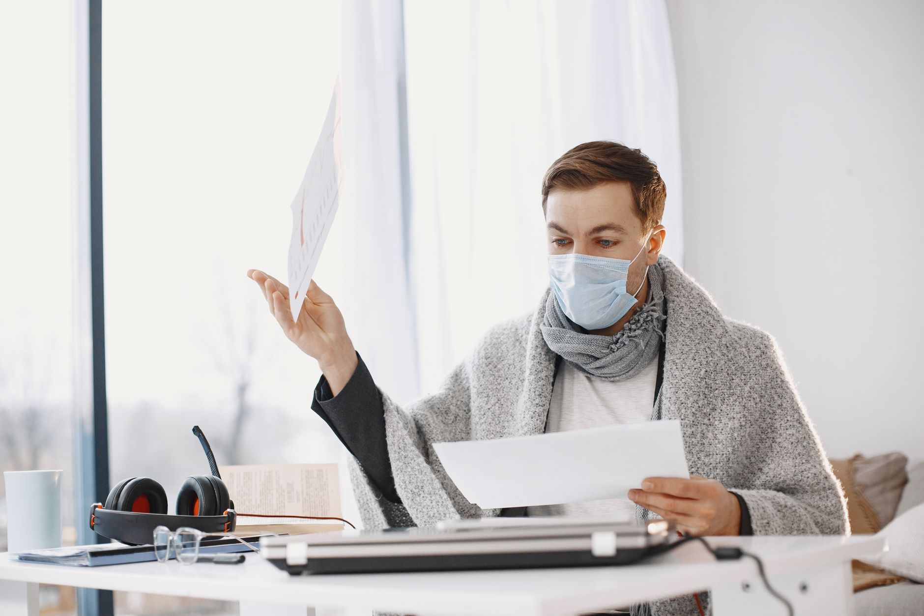 Man wearing protective mask working from home, wrapped in a blanket, illustrating remote work during illness.