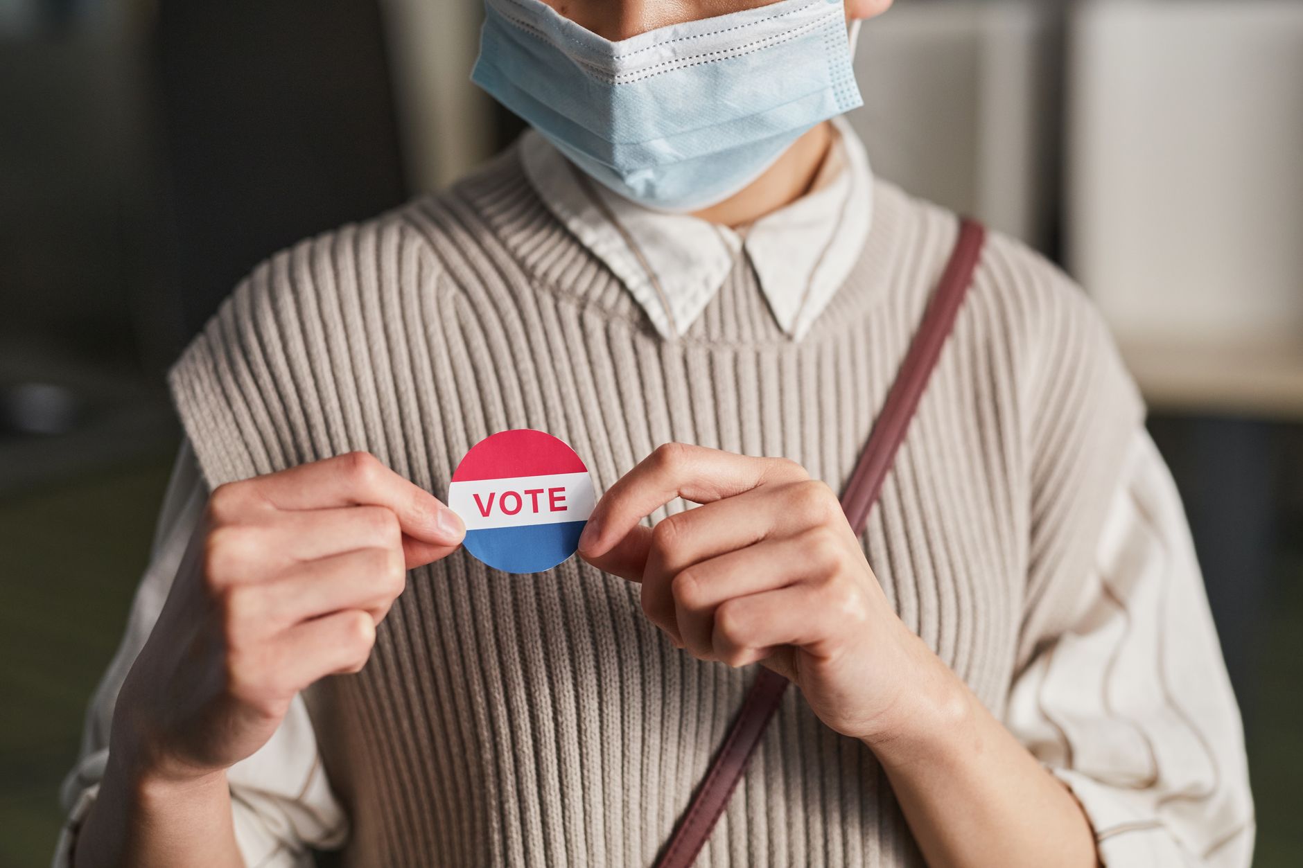 A person wearing a mask holds a voting badge, symbolizing participation in the election.