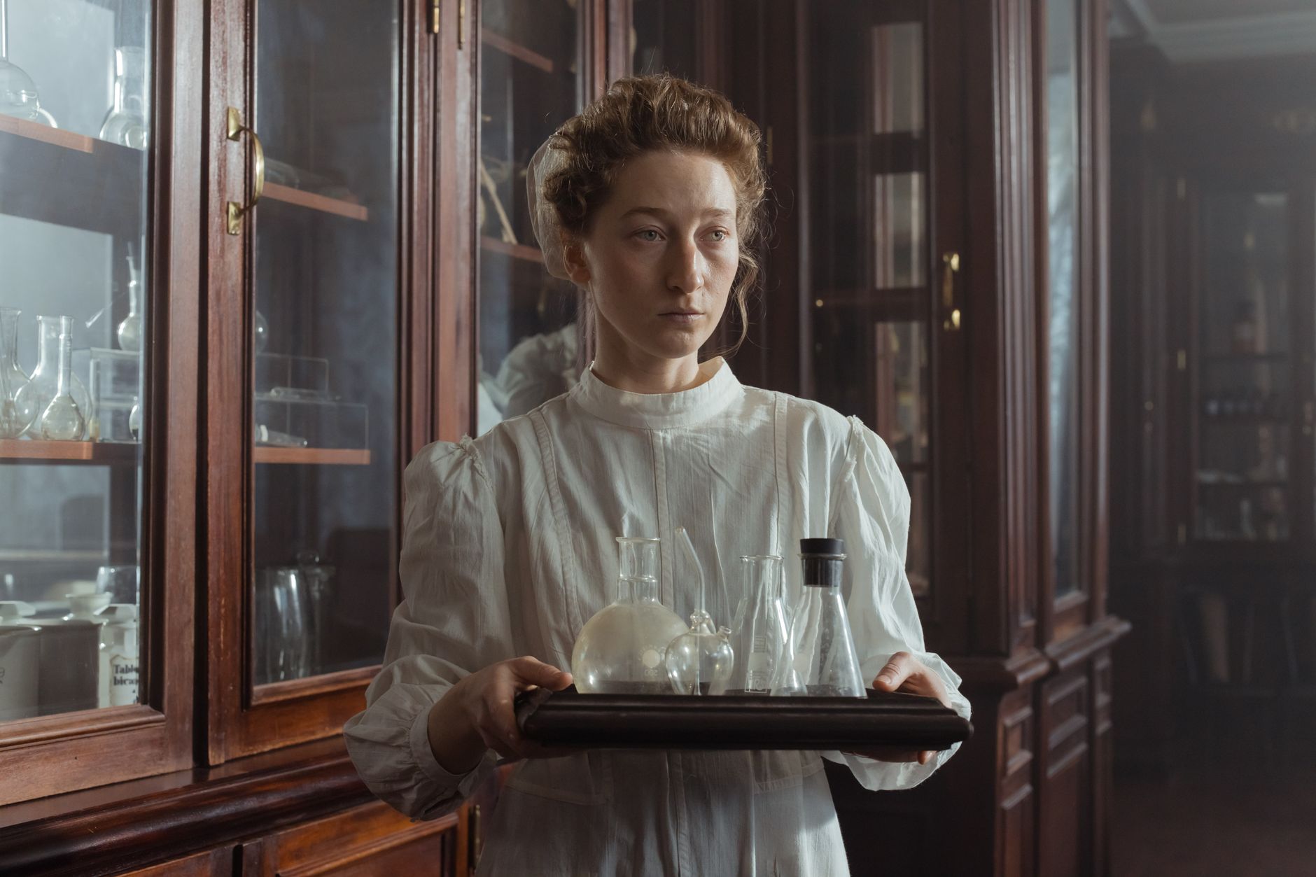 A woman in a vintage laboratory holding a tray with glass flasks and bottles, showcasing pharmaceutical history.