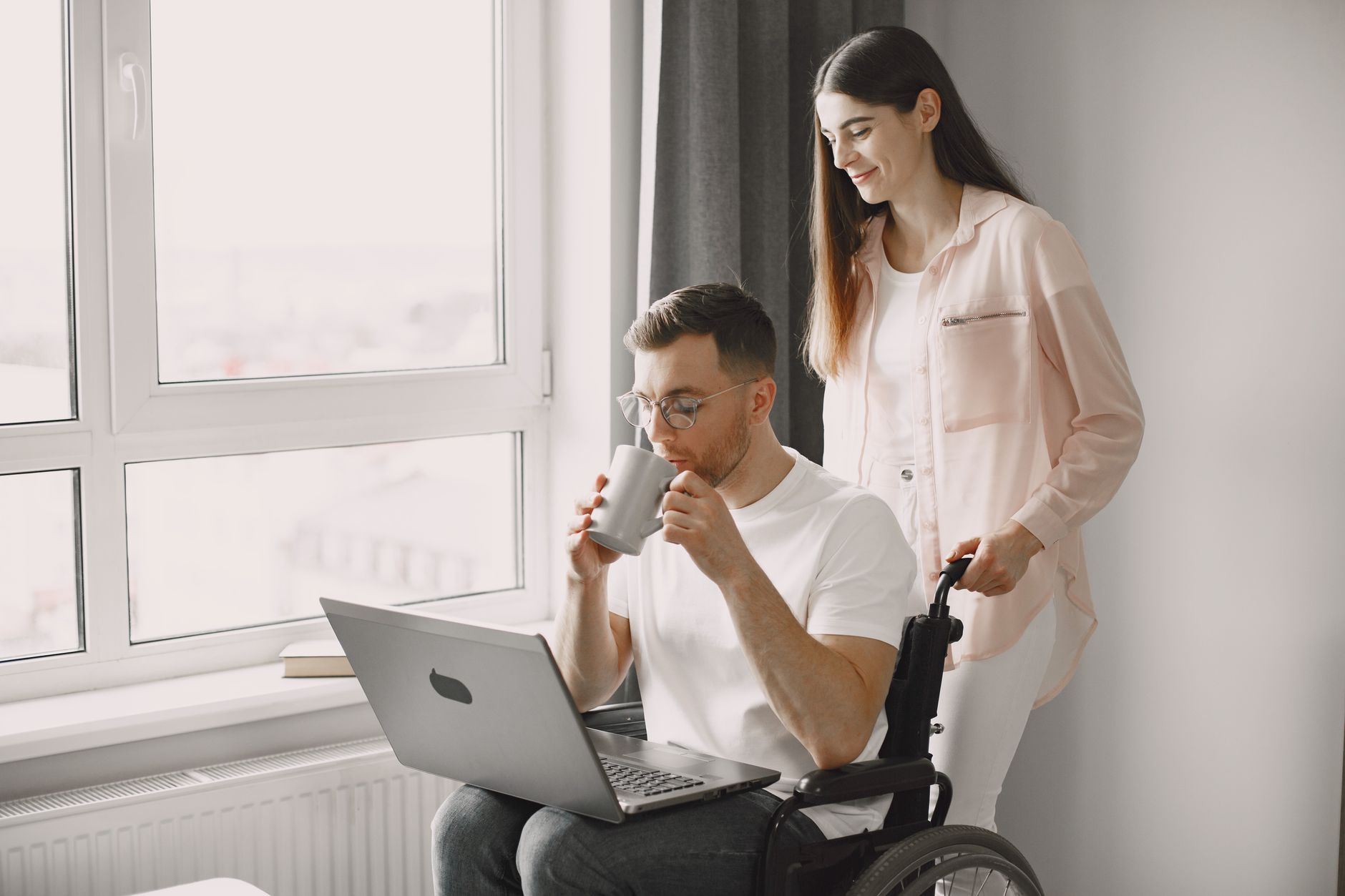 A caring woman assists a man in a wheelchair as they enjoy coffee and work on a laptop indoors.