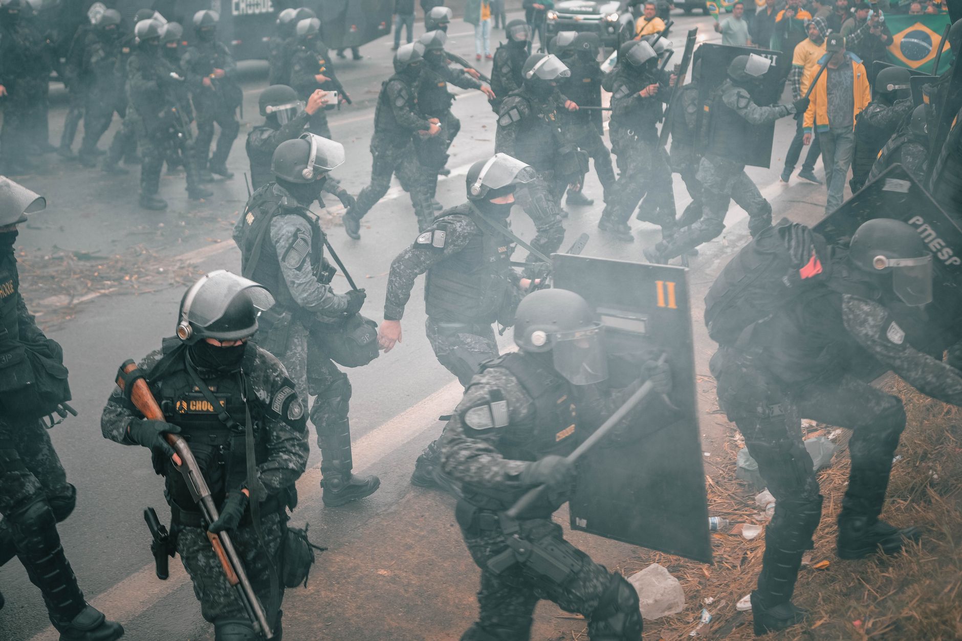 Riot police with shields confront protesters in a smoky urban setting.